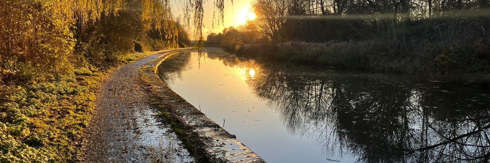 low bright sun reflecting trees in a canal bordered by wide muddy towpath and stone edge shining in the sun. Credit Helen Clayton